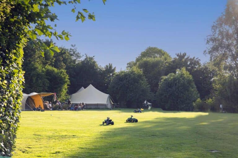 Sfeervolle, groene camping met ruime glamping tenten en speelgoedtractors op een groot, zonnig grasveld in de natuur.